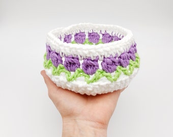 May include: A white crocheted bowl with a purple and green floral design. The bowl is handmade and has a delicate, intricate pattern.