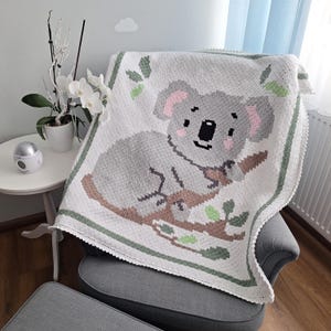 Puede incluir: Manta blanca de ganchillo con diseño de koala gris, orejas rosas, sentado en una rama marrón con hojas verdes. Borde verde y blanco, sobre un sillón gris.