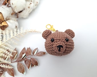 May include: A brown crocheted bear head keyring with black eyes and a black nose. The bear has a gold-coloured clasp for attaching to keys or bags. The bear is a handmade item.