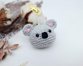 May include: A crocheted koala keychain with a gold-colored clasp. The koala is gray with pink accents on the ears and cheeks, and black eyes and nose. The keychain is a handmade item.