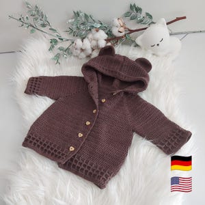 May include: A brown, hooded baby sweater with bear ears. The sweater has a button-up front with wooden heart-shaped buttons. The garment is crocheted and has a textured pattern. The sweater is displayed on a white, fluffy surface.