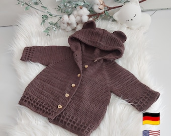 May include: A brown, hooded baby sweater with bear ears. The sweater has a button-up front with wooden heart-shaped buttons. The garment is crocheted and has a textured pattern. The sweater is displayed on a white, fluffy surface.
