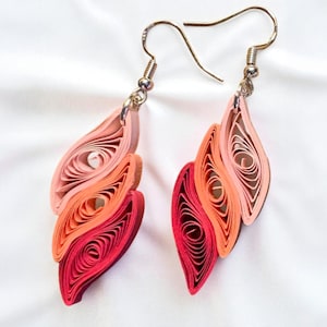 May include: A pair of handmade paper quilled earrings. The earrings feature a layered design in shades of pink, orange, and red. The earrings have silver-coloured ear wires and are displayed against a white background.
