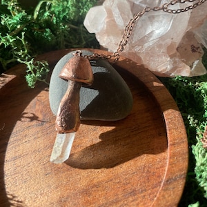 Puede incluir: Un collar con un colgante de hongo de cobre y un tallo de cristal de cuarzo transparente. El collar se muestra sobre una superficie de madera con musgo y un grupo de cristales de cuarzo transparentes.
