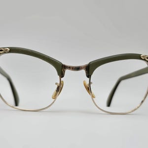 May include: Olive green and gold-tone eyeglasses with a cat-eye shape. The frames have a decorative gold floral design on the upper corners. The lenses are clear, and the nose pads are a light tan color.