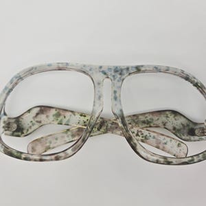 May include: A pair of clear, oversized sunglasses with a mottled, speckled pattern in shades of green, blue, and pink.