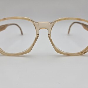 May include: A pair of spectacles with a light brown, oval-shaped frame. The lenses are clear, and the arms are curved. The spectacles have a classic, vintage design. The frame is made of a light-coloured material.