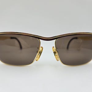 May include: Gold-framed sunglasses with brown lenses. The sunglasses have a rectangular shape with a curved top bar and a gold bridge. The temples are thin and gold, and the lenses are a solid brown color.