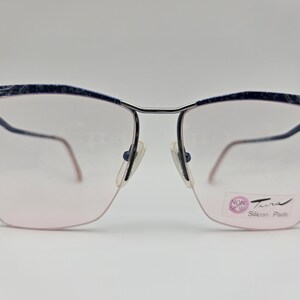 May include: Vintage eyeglasses with a silver metal frame and blue patterned top rims. The lenses are a light pink color. The glasses have a rectangular shape with rounded edges. The brand name "Turra" is visible on the lens.