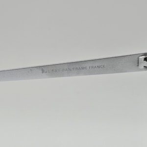 May include: Close-up of the silver metal frame of a pair of Ray-Ban sunglasses. The frame has the text "Ray-Ban/Frame France" engraved on it.