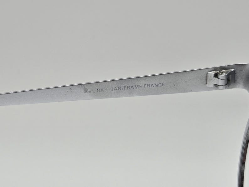 May include: Close-up of the silver metal frame of a pair of Ray-Ban sunglasses. The frame has the text "Ray-Ban/Frame France" engraved on it.