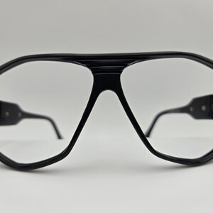 May include: Black plastic framed glasses with gold accents. The glasses have a geometric shape and clear lenses.