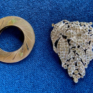 May include: A gold-toned metal ring with a decorative design and a silver-toned brooch with a floral design featuring rhinestones.