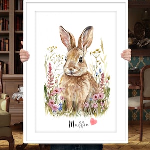 Puede incluir: Una pintura de acuarela de un conejo marr&oacute;n sentado en un campo de flores silvestres. El conejo est&aacute; mirando directamente al espectador. Las flores son rosas, azules y amarillas. El texto "Muffin" est&aacute; escrito debajo del conejo, con un coraz&oacute;n rosa al lado.