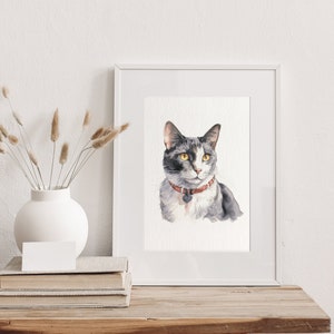 Puede incluir: Una pintura de acuarela de un gato gris y blanco con ojos amarillos que lleva un collar rojo con una etiqueta. La pintura est&aacute; enmarcada en un marco blanco y se muestra en una mesa de madera.