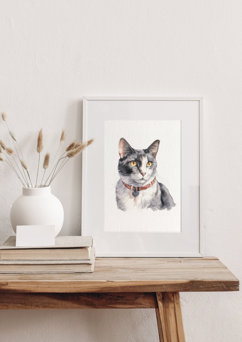 Puede incluir: Una pintura de acuarela de un gato gris y blanco con ojos amarillos que lleva un collar rojo con una etiqueta. La pintura est&aacute; enmarcada en un marco blanco y se muestra en una mesa de madera.