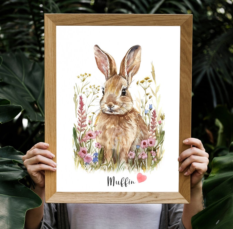 Puede incluir: Una pintura de acuarela de un conejo marr&oacute;n sentado en un campo de flores rosas y azules. El conejo est&aacute; mirando al espectador y tiene un coraz&oacute;n rosa sobre su cabeza. El texto "Muffin" est&aacute; escrito debajo del conejo.