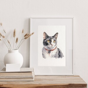 May include: A watercolor painting of a gray and white cat with yellow eyes wearing a red collar with a tag. The painting is framed in a white frame and is displayed on a wooden table.
