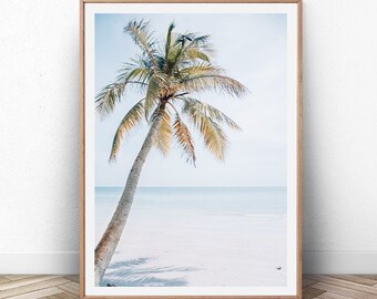 May include: A single palm tree stands on a white sandy beach with blue water in the background. The sky is a light blue with white clouds. The image is framed in a light wood frame.