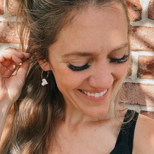 May include: A woman with long blonde hair pulled back in a ponytail is wearing a black tank top and gold hoop earrings with a small white heart charm. She is smiling and looking down.
