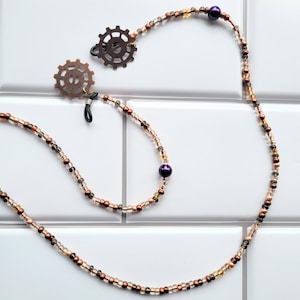 May include: A beaded eyeglass chain featuring a mix of small copper, bronze, and clear glass beads interspersed with larger, smooth purple beads. The chain attaches to eyewear with two bronze-toned metal gear-shaped charms and black rubber loops.