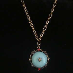 May include: A gold chain necklace with a light blue pendant. The pendant is round and has a decorative border with red and white stones.