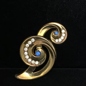 May include: A gold-toned vintage brooch with a swirling design. The brooch features clear and blue rhinestones. The design includes two spirals and a curved base. The background is black.