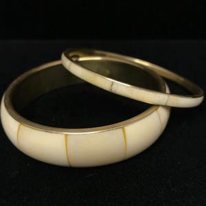 Puede incluir: Dos brazaletes de color marfil con detalles met&aacute;licos dorados. El brazalete m&aacute;s grande tiene rayas amarillas verticales. El m&aacute;s peque&ntilde;o descansa sobre el m&aacute;s grande. El fondo es negro.
