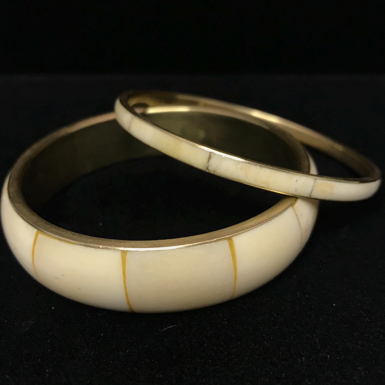 Puede incluir: Dos brazaletes de color marfil con detalles met&aacute;licos dorados. El brazalete m&aacute;s grande tiene rayas amarillas verticales. El m&aacute;s peque&ntilde;o descansa sobre el m&aacute;s grande. El fondo es negro.