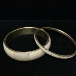 Puede incluir: Dos brazaletes de color marfil con bandas met&aacute;licas de color lat&oacute;n. El brazalete m&aacute;s grande tiene una banda m&aacute;s ancha con incrustaciones rectangulares de color marfil. El brazalete m&aacute;s peque&ntilde;o es m&aacute;s delgado con incrustaciones similares.