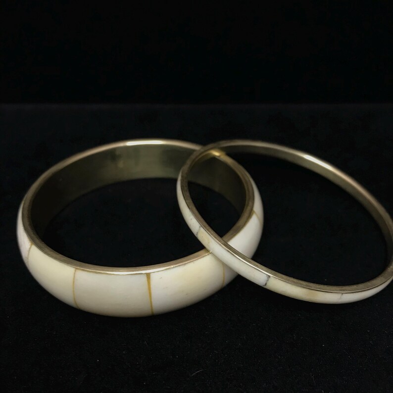 Puede incluir: Dos brazaletes de color marfil con bandas met&aacute;licas de color lat&oacute;n. El brazalete m&aacute;s grande tiene una banda m&aacute;s ancha con incrustaciones rectangulares de color marfil. El brazalete m&aacute;s peque&ntilde;o es m&aacute;s delgado con incrustaciones similares.
