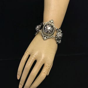 Puede incluir: Un brazalete de plata con un gran cabujón redondo con estampado blanco y negro. El brazalete tiene una orfebrería adornada y detallada.