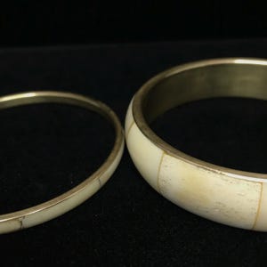 Puede incluir: Dos brazaletes de color marfil con bandas de metal dorado. El brazalete m&aacute;s grande tiene una banda m&aacute;s ancha con incrustaciones rectangulares de color marfil. El brazalete m&aacute;s peque&ntilde;o es una banda delgada con una superficie lisa.