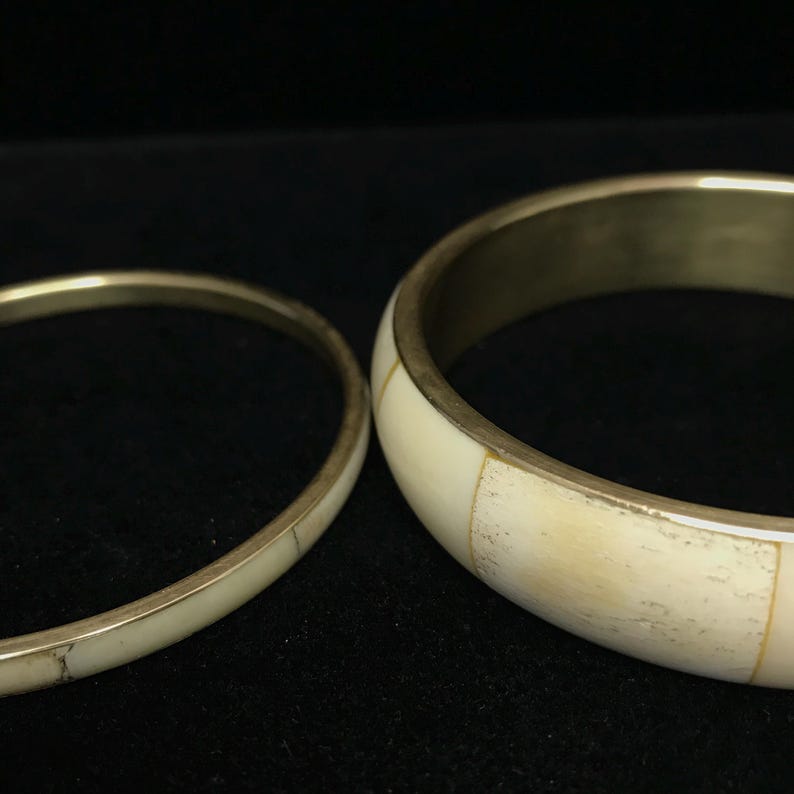 Puede incluir: Dos brazaletes de color marfil con bandas de metal dorado. El brazalete m&aacute;s grande tiene una banda m&aacute;s ancha con incrustaciones rectangulares de color marfil. El brazalete m&aacute;s peque&ntilde;o es una banda delgada con una superficie lisa.