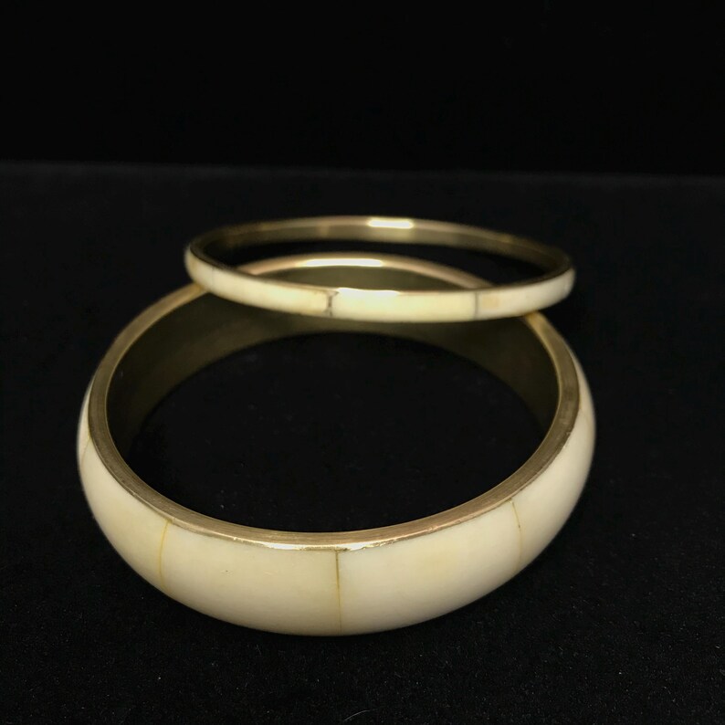 Puede incluir: Dos brazaletes de color marfil con detalles met&aacute;licos dorados. El brazalete m&aacute;s grande es m&aacute;s ancho y tiene forma curva, mientras que el m&aacute;s peque&ntilde;o es m&aacute;s delgado y se encuentra encima. Los brazaletes probablemente est&aacute;n hechos de un material compuesto.
