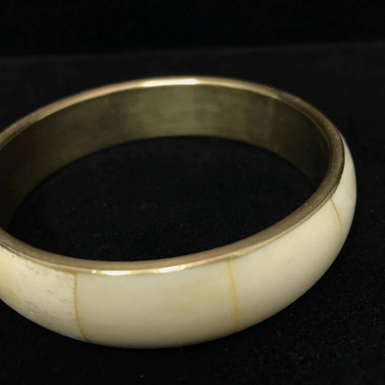 Puede incluir: Una pulsera de brazalete dorada con una incrustaci&oacute;n de color crema. La pulsera tiene un dise&ntilde;o liso y redondeado con un acabado pulido. Las secciones de color crema est&aacute;n separadas por finas l&iacute;neas doradas. Es una joya.