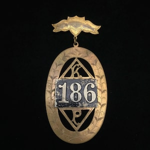 May include: A gold-colored metal pendant with a black and white enamel plaque featuring the number "186". The pendant is shaped like an oval with a diamond-shaped cutout in the center. The pendant is attached to a gold-colored metal chain with a decorative top.