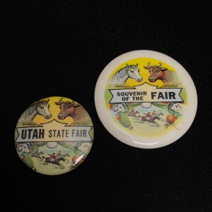 May include: Two vintage Utah State Fair souvenir pinback buttons. One button features a horse and a cow with the text "Utah State Fair". The other button says "Souvenir of the Fair" with a horse and cow image.