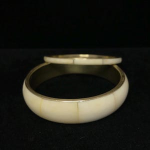 Puede incluir: Dos brazaletes de color marfil con detalles met&aacute;licos dorados. El brazalete m&aacute;s grande es m&aacute;s ancho, mientras que el m&aacute;s peque&ntilde;o es m&aacute;s delgado. Los brazaletes est&aacute;n sobre un fondo oscuro, resaltando su color y dise&ntilde;o. Joyer&iacute;a.