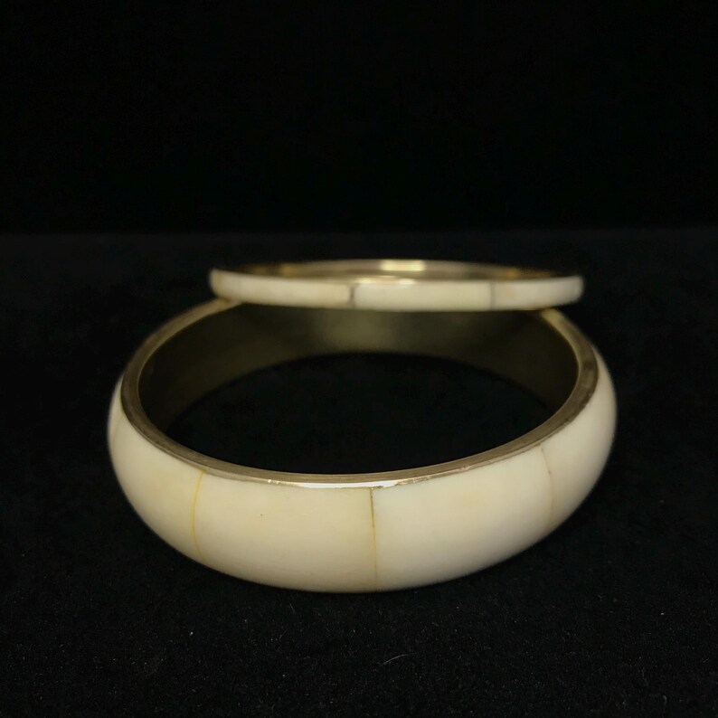 Puede incluir: Dos brazaletes de color marfil con detalles met&aacute;licos dorados. El brazalete m&aacute;s grande es m&aacute;s ancho, mientras que el m&aacute;s peque&ntilde;o es m&aacute;s delgado. Los brazaletes est&aacute;n sobre un fondo oscuro, resaltando su color y dise&ntilde;o. Joyer&iacute;a.