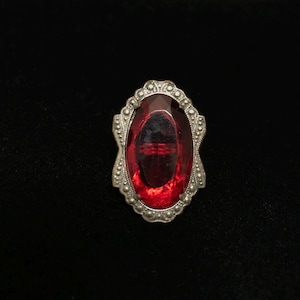 Op de afbeelding: Een vintage broche met een grote, ovale, rode glazen steen gezet in een zilverkleurig metalen frame met een decoratief ontwerp.