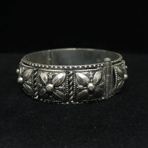 May include: A silver-toned hinged bracelet with a floral design. The bracelet features a series of embossed flowers and beaded accents. The clasp is visible on the side. The bracelet is set against a dark background.