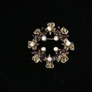 Puede incluir: Un broche dorado con un diseño circular que presenta piedras de imitación rojas y transparentes. El broche está engastado en un patrón de filigrana.