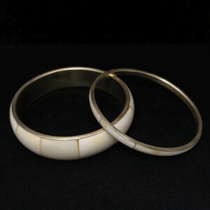 Puede incluir: Dos brazaletes de color marfil con bordes de metal plateado. El brazalete m&aacute;s ancho est&aacute; segmentado con piezas rectangulares de marfil. El brazalete m&aacute;s delgado es una banda s&oacute;lida con detalles en marfil. Sobre fondo negro.