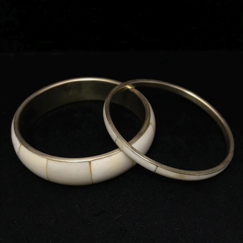 Puede incluir: Dos brazaletes de color marfil con bordes de metal plateado. El brazalete m&aacute;s ancho est&aacute; segmentado con piezas rectangulares de marfil. El brazalete m&aacute;s delgado es una banda s&oacute;lida con detalles en marfil. Sobre fondo negro.