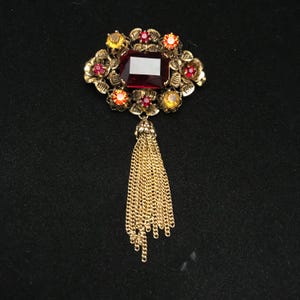 May include: A vintage gold-toned brooch with a large, rectangular, red gemstone at its center. The brooch is adorned with smaller red, orange, and yellow gemstones and floral accents. A gold chain tassel hangs from the bottom, adding a touch of elegance.