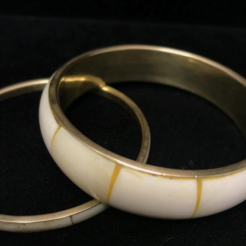 Puede incluir: Dos brazaletes de color marfil con bordes de metal dorado y detalles amarillos. Los brazaletes est&aacute;n apilados, uno parcialmente oscureciendo al otro. El fondo es negro.