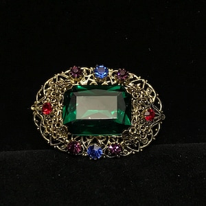 May include: An oval-shaped brooch with a large emerald-green rectangular center stone. The brooch is framed by a gold filigree design and accented with red, blue, and purple gemstones. The background is black.