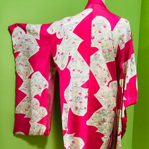 May include: A vibrant pink kimono with a floral pattern in cream and pastel colors. The kimono is displayed on a mannequin, showcasing its flowing sleeves and intricate design. The floral pattern is repeated throughout the garment.