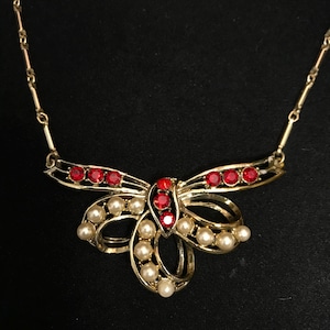 May include: A gold-tone necklace featuring a central bow-shaped pendant. The pendant is decorated with small white faux pearls and bright red round rhinestones. The chain consists of thin gold-tone metal bars connected by small circular links.